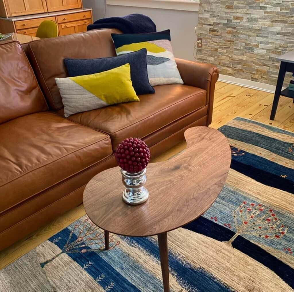 A cozy living room features a comfortable brown leather sofa adorned with colorful decorative pillows, a unique kidney-shaped walnut coffee table by stevecastleswoodworker/SCWoodworker with a red berry centerpiece, and a striped blue and beige area rug with tree motifs, creating an inviting and stylish interior design.