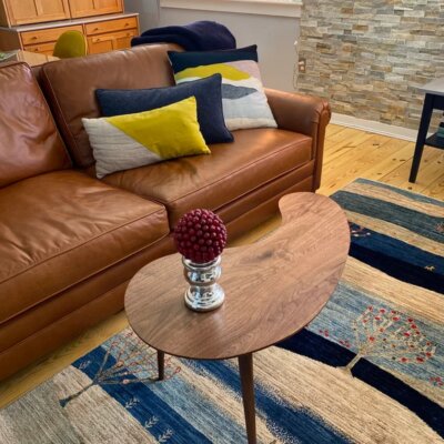 A cozy living room features a comfortable brown leather sofa adorned with colorful decorative pillows, a unique kidney-shaped walnut coffee table by stevecastleswoodworker/SCWoodworker with a red berry centerpiece, and a striped blue and beige area rug with tree motifs, creating an inviting and stylish interior design.