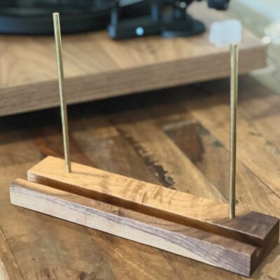 A handcrafted wooden display stand featuring two vertical brass rods, designed to hold vinyl records, resting on a rustic wooden table with a blurred turntable in the background.